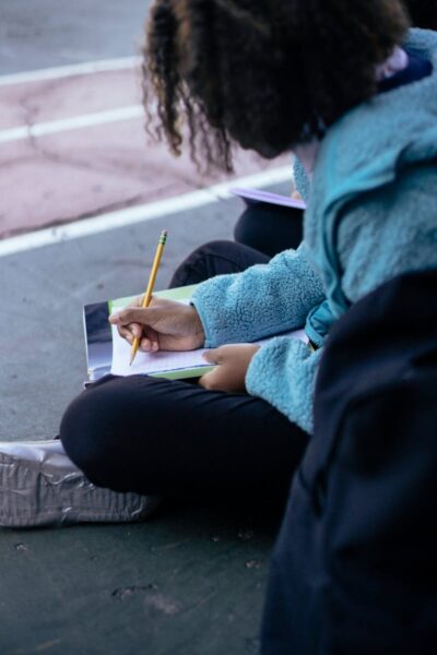 A young girl writing with a pencil while sitting outdoors in casual attire.
